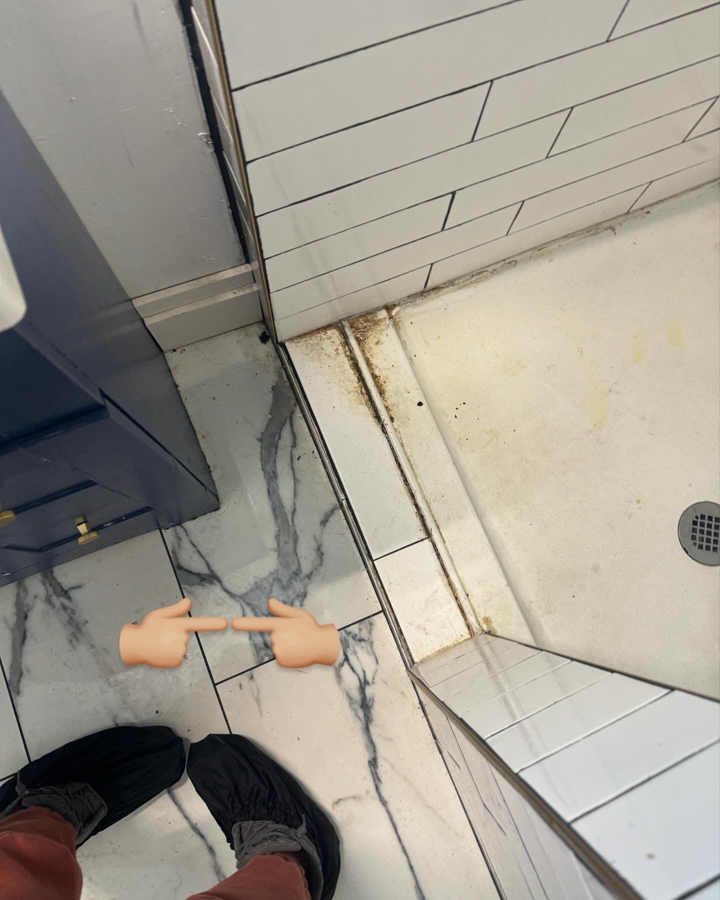 Person's feet and hands pointing to rusty mold in a shower corner with white tiles and dark blue cabinet.
