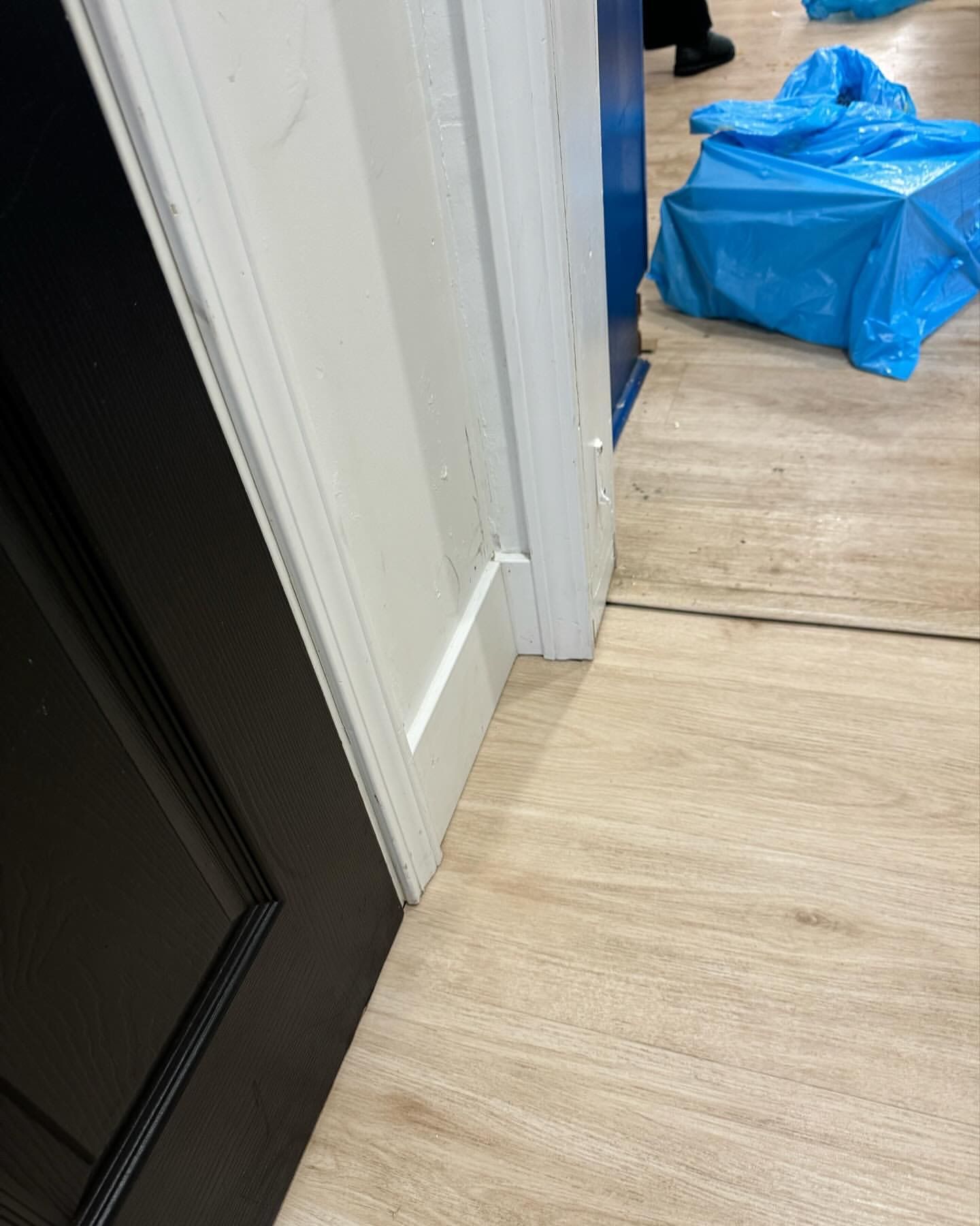 White door frame slightly warped next to a black door; light wood-look floor; blue protective covering in the background.
