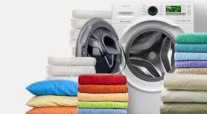 Washing machine with open door and stacks of folded towels, sheets, and pillows in various colors.