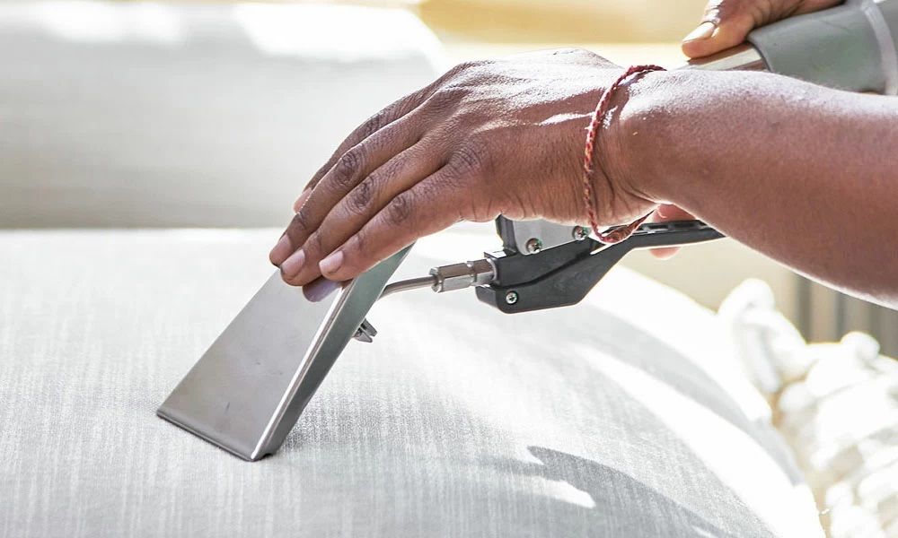 A person using a upholstery cleaner on a white couch, close-up of hands and tool.