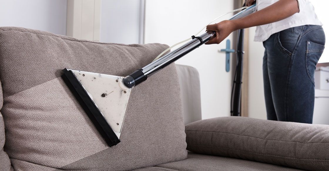 Person vacuuming a beige couch with a handheld attachment, indoors.