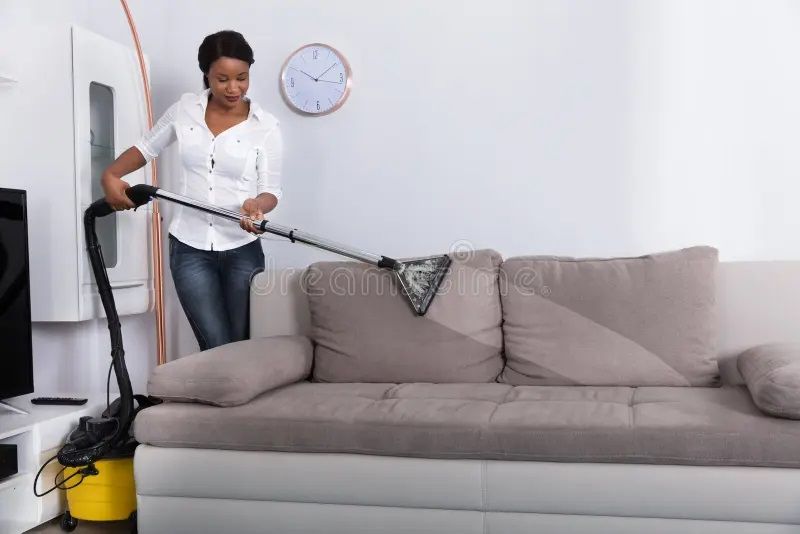 Woman vacuuming a gray sofa in a living room.