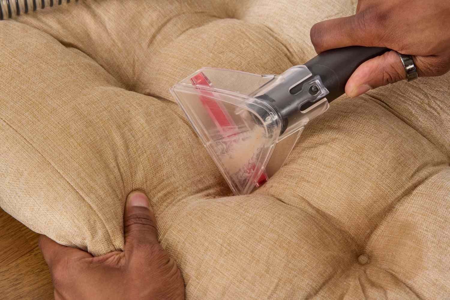 Person cleaning a tan upholstered cushion with a handheld upholstery cleaner.