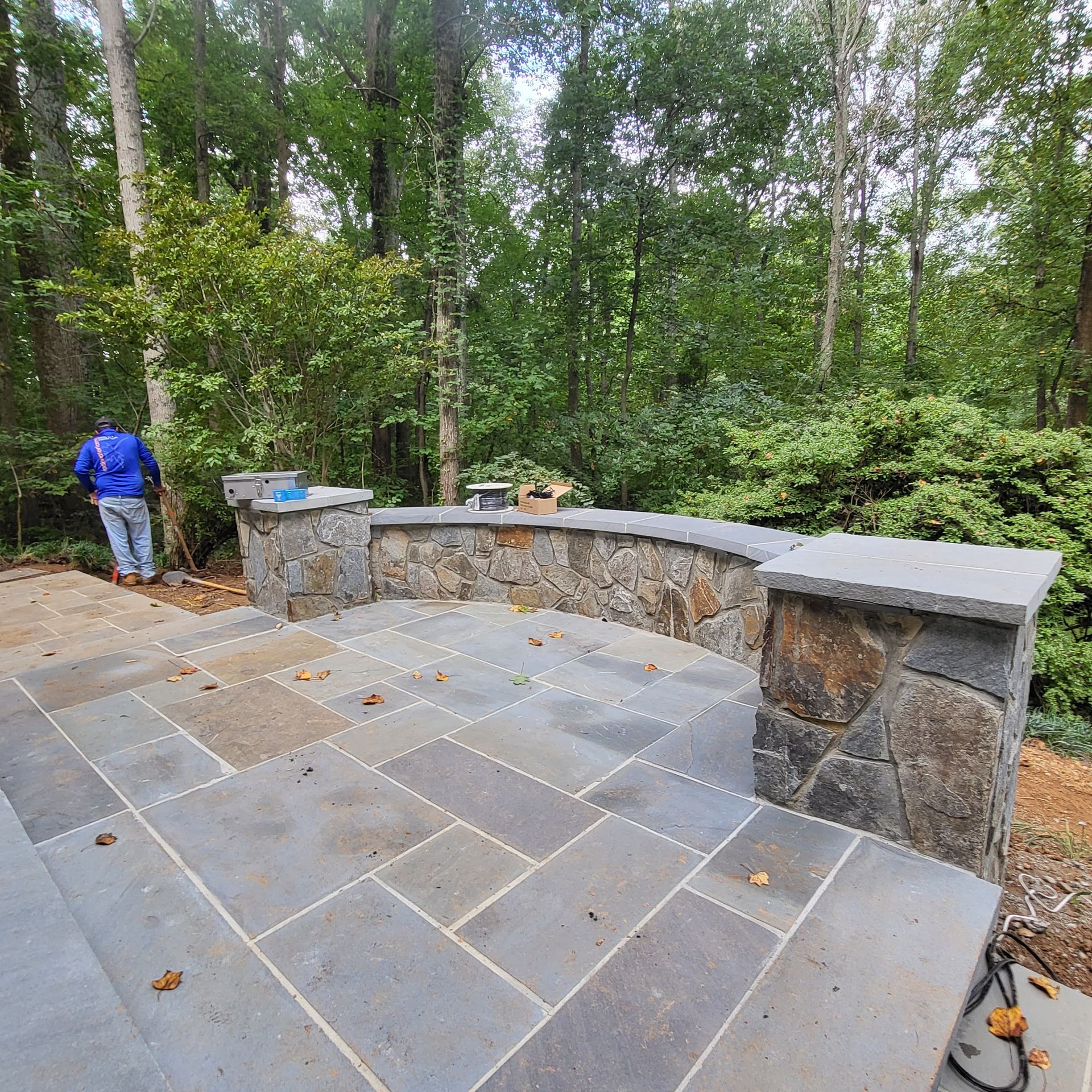 A man in a blue jacket is standing on a stone patio in the woods.