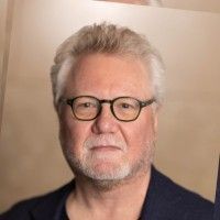 A man wearing glasses and a blue suit is standing in front of a white background.