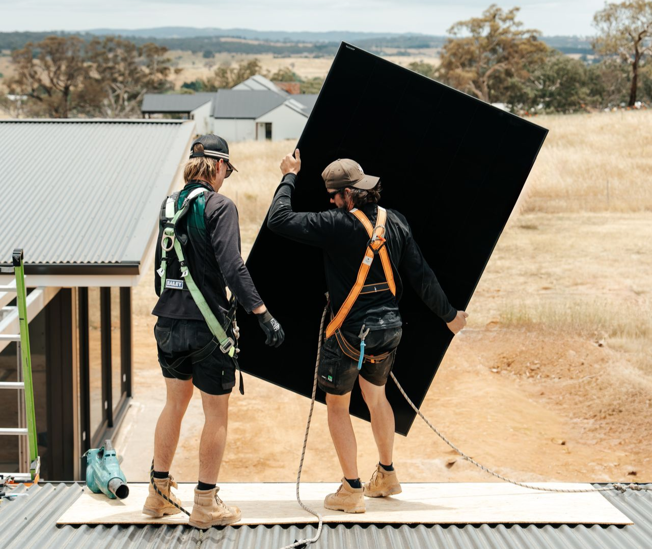 Two workers installing a solar panel on a rooftop. They are wearing safety harnesses and work boots.