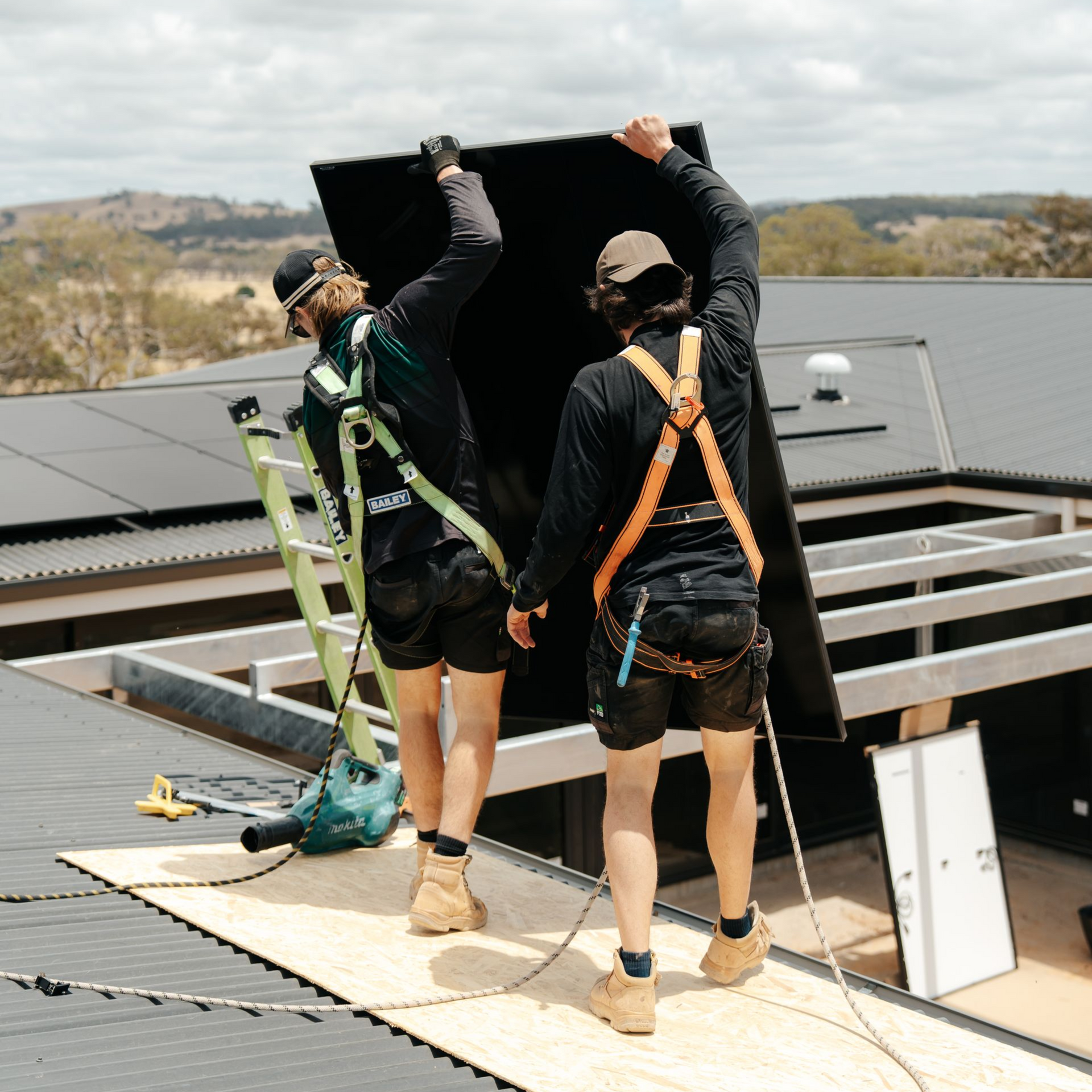 Two workers installing a solar panel on a rooftop. They are wearing safety harnesses and work boots.