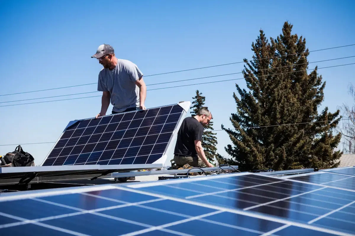 Two workers in casual clothing install solar panels on a sunny rooftop under a clear blue sky.