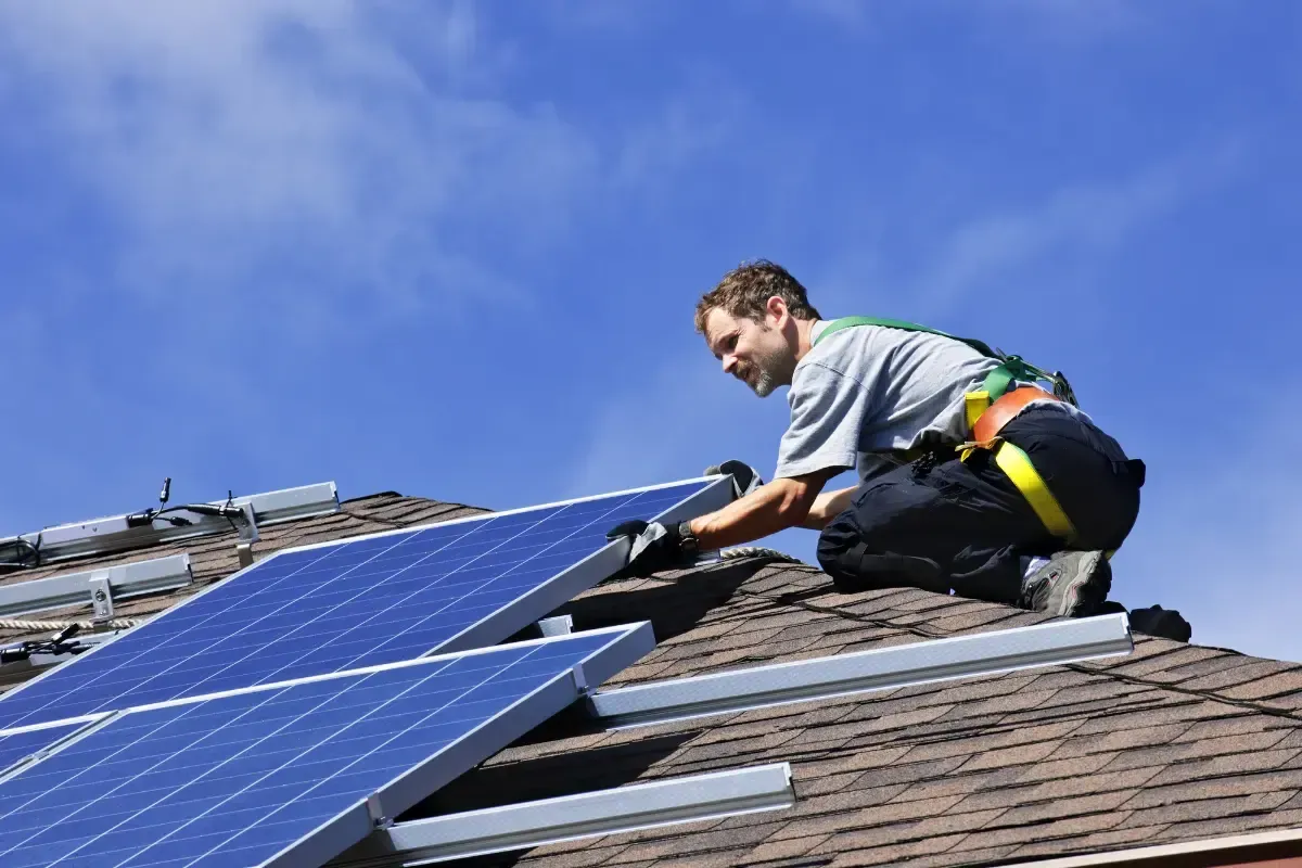 A person wearing safety gear kneeling on a roof, installing solar panels under a clear blue sky.