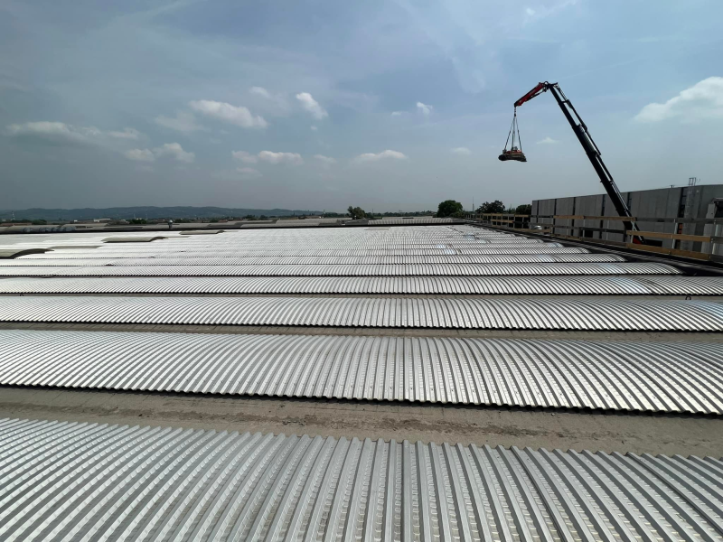 Una gru solleva un carico sopra un grande tetto caratterizzato da file parallele di pannelli metallici ondulati di colore argento.