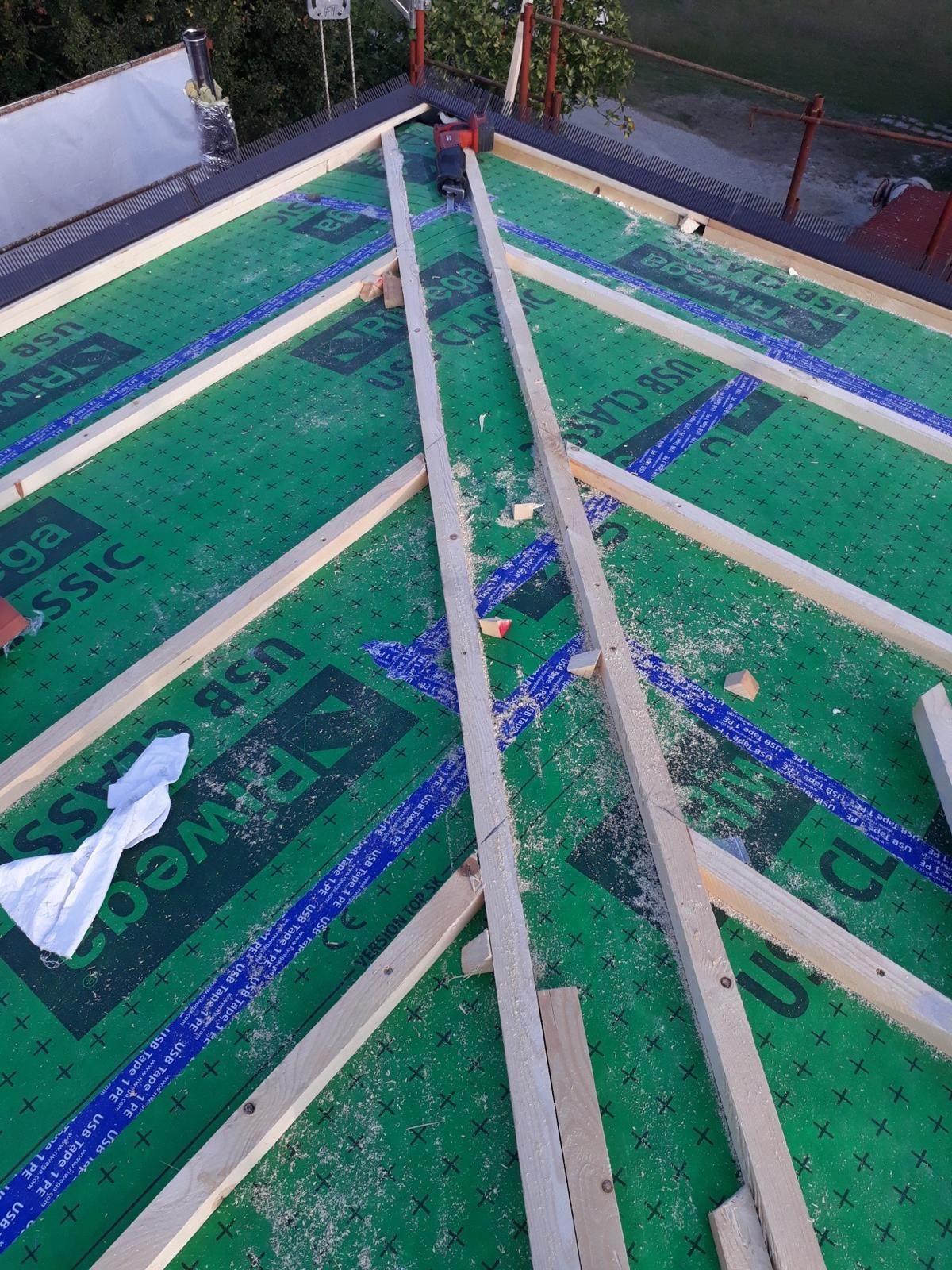 Una vista dall'alto di un tetto in costruzione, che mostra le travi di supporto in legno installate sopra una membrana impermeabile verde.