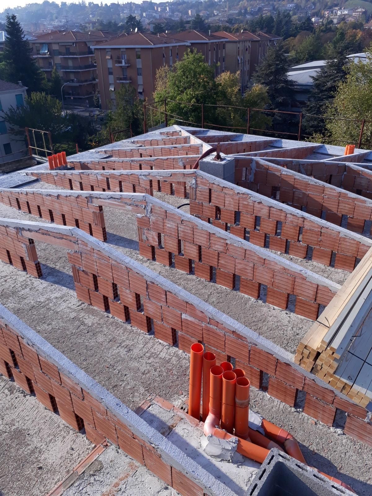 Vista dall'alto del tetto di un edificio in costruzione, con pareti in mattoni arancioni, strutture di supporto e tubature.