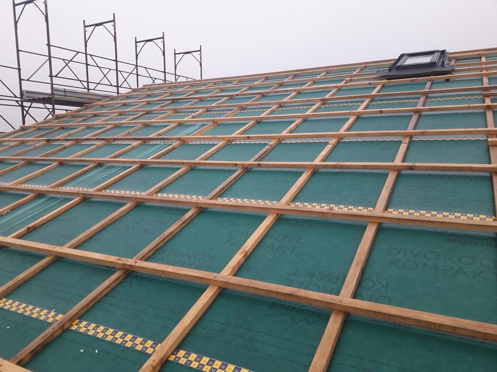 Un tetto a falde in costruzione, caratterizzato da una struttura a griglia di listelli di legno sopra una membrana impermeabile verde e un lucernario.