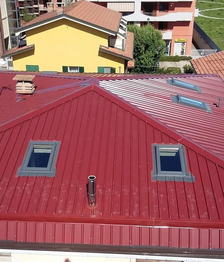 Un tetto in metallo con pannelli verticali rossi, caratterizzato da due lucernari, un camino e uno sfondo di edifici del quartiere.