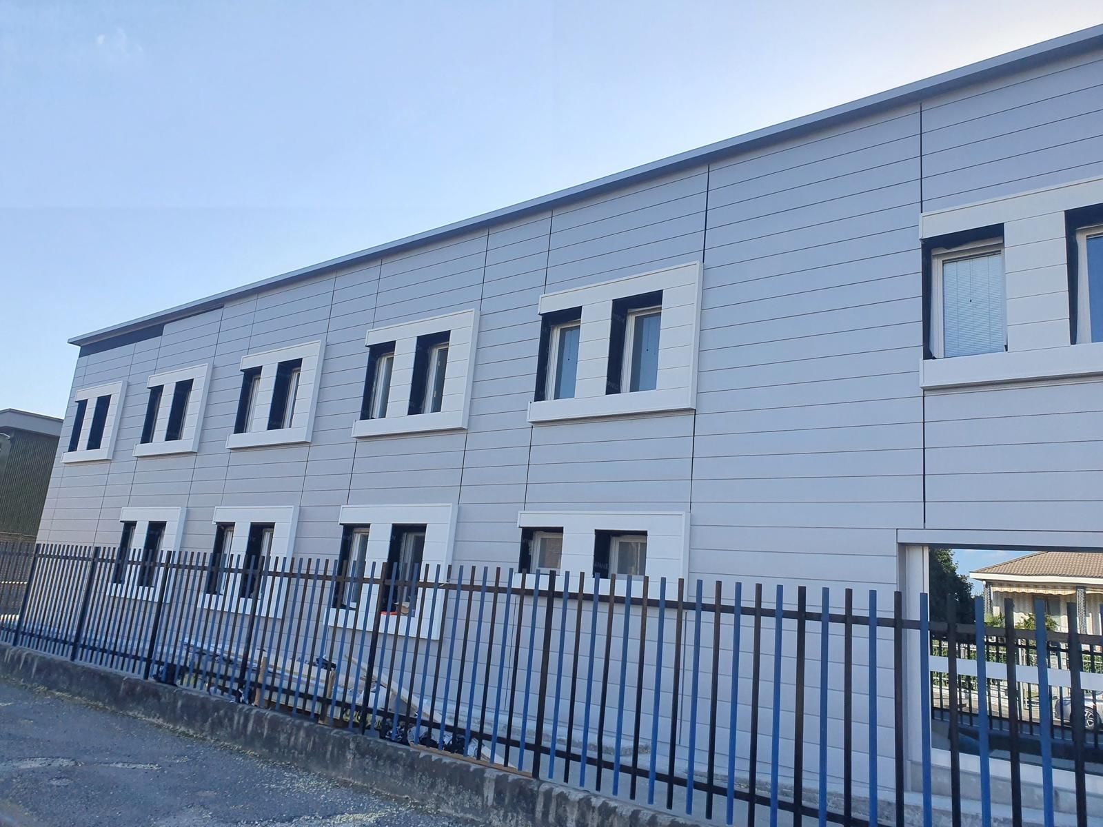 Un edificio a due piani, con rivestimento esterno in metallo grigio e finestre con cornici bianche, visto da dietro una recinzione metallica nera in una giornata limpida.