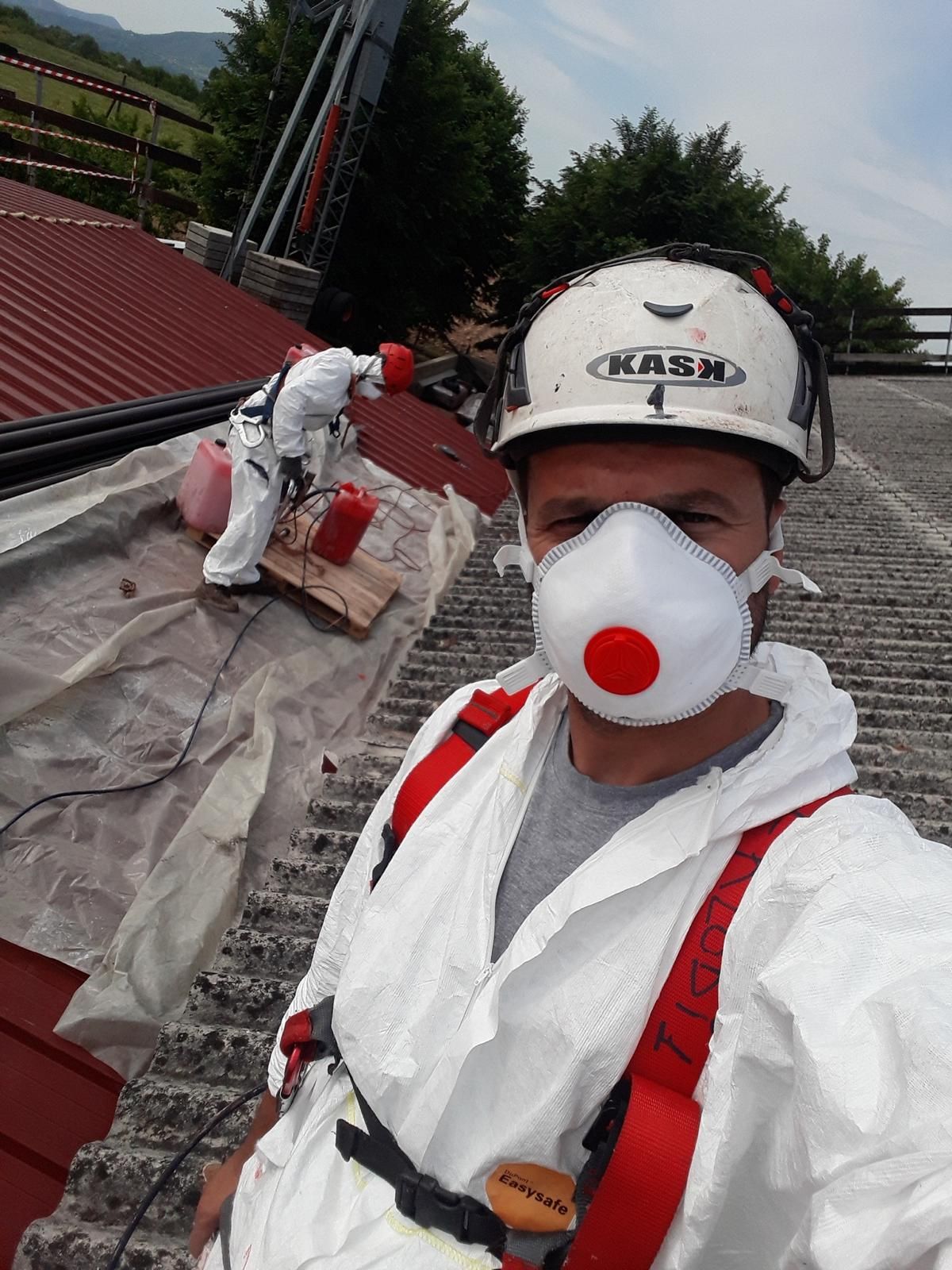 Una persona con indosso un elmetto, una maschera protettiva e una tuta bianca si scatta un selfie su un tetto, mentre un'altra persona lavora alle sue spalle.