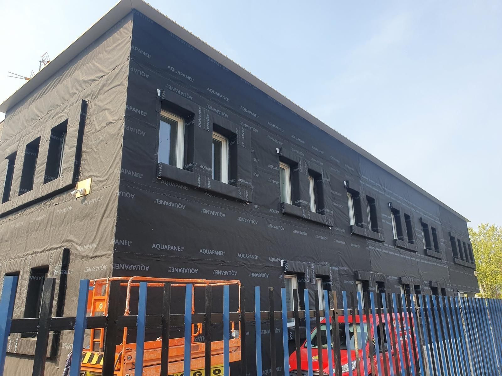 Un edificio a due piani in costruzione, ricoperto da un telo nero resistente alle intemperie, visibile dietro una recinzione metallica.