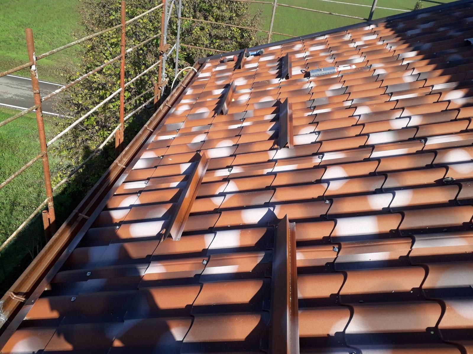 Un primo piano di un tetto di tegole marroni in costruzione, che mostra paraneve metallici parzialmente installati e impalcature nelle vicinanze.