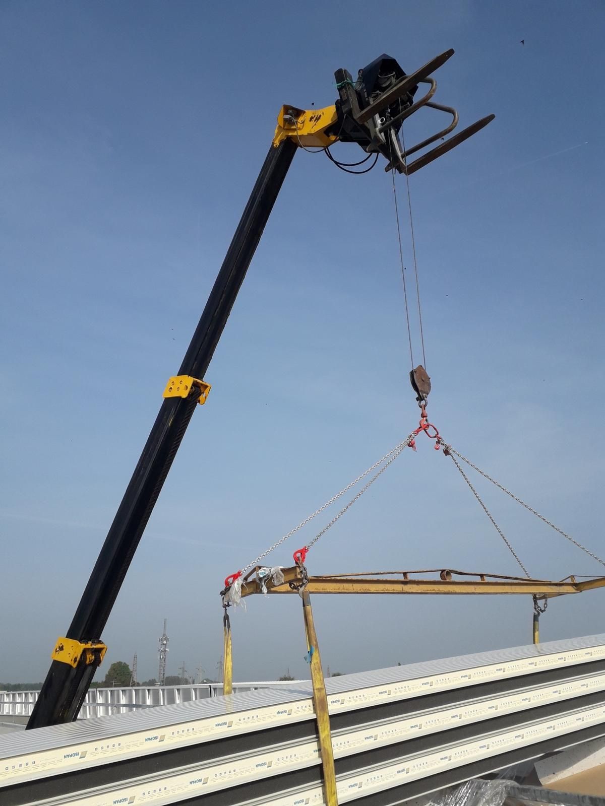 Un sollevatore telescopico con un braccio giallo solleva un lungo pannello edile con delle cinghie, sullo sfondo di un cielo azzurro e limpido.