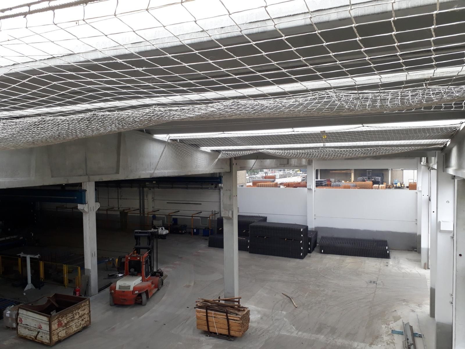 Interno di un magazzino con soffitto a rete bianca, un carrello elevatore rosso parcheggiato e pile di materiali scuri rettangolari.