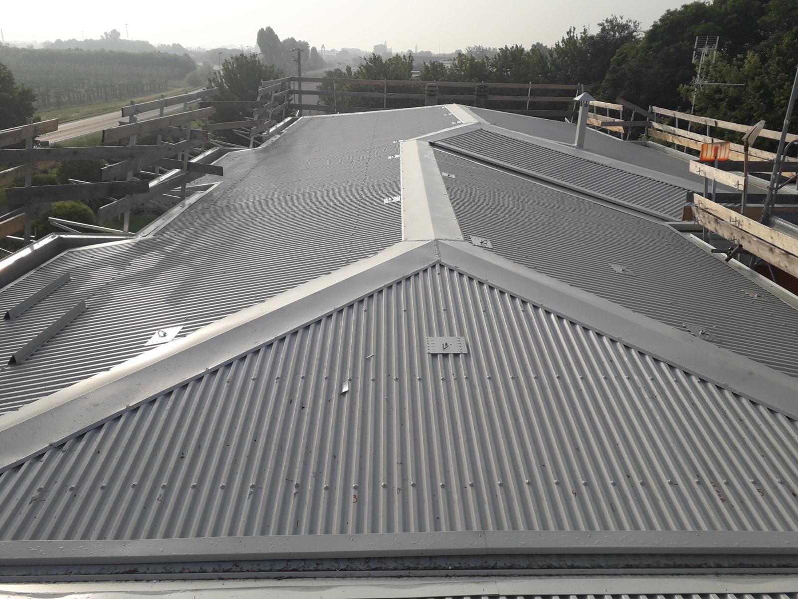Vista dall'alto di un tetto in lamiera ondulata grigia con diverse calotte di colmo e sezioni in costruzione.