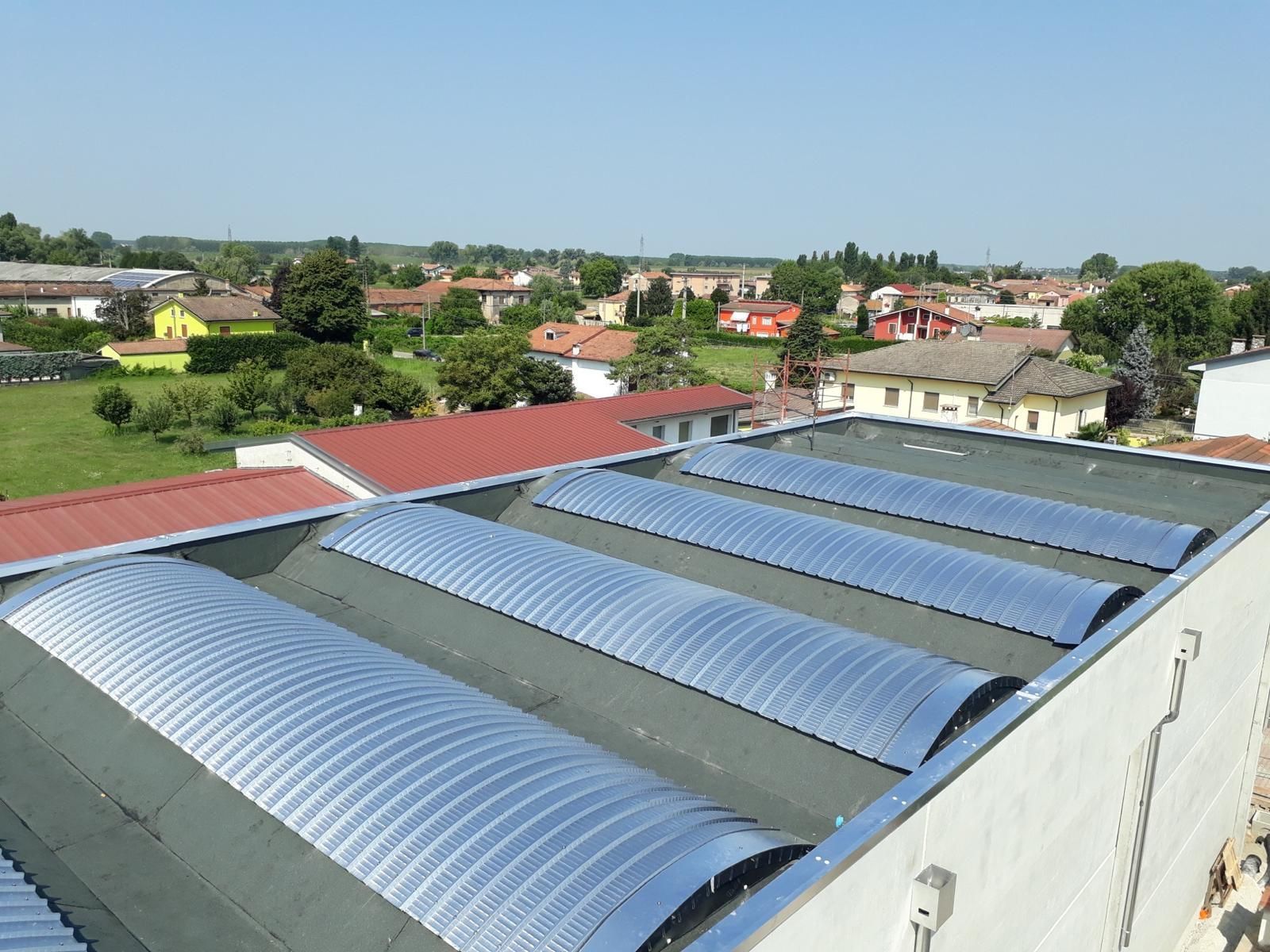 Vista dall'alto di un tetto piano caratterizzato da diversi lucernari paralleli, curvi e traslucidi che si affacciano su un paesaggio rurale.