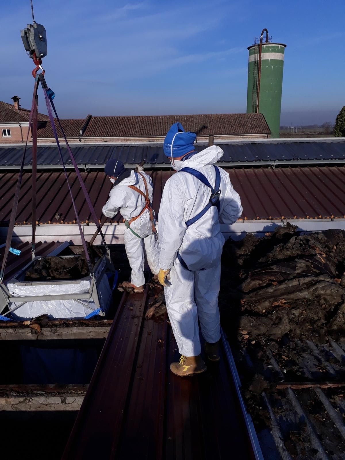 Due operai con tute protettive bianche e imbracature di sicurezza lavorano su un tetto industriale, utilizzando una gru per rimuovere i detriti.