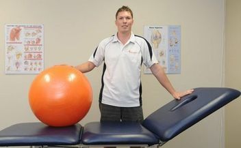A man is standing next to an orange exercise ball on a table.