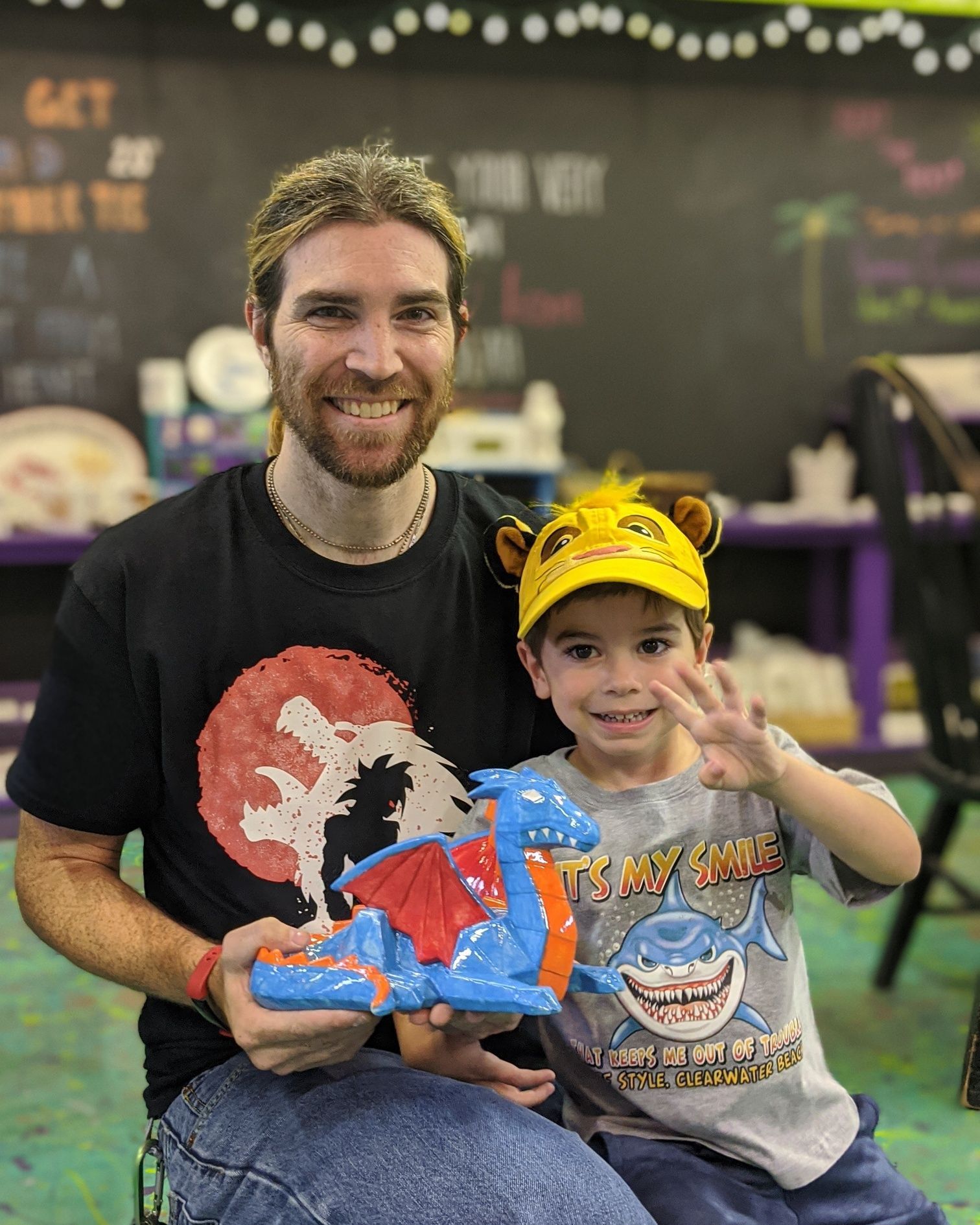 Man and child smiling, holding a painted blue dragon with red wings, inside a studio.