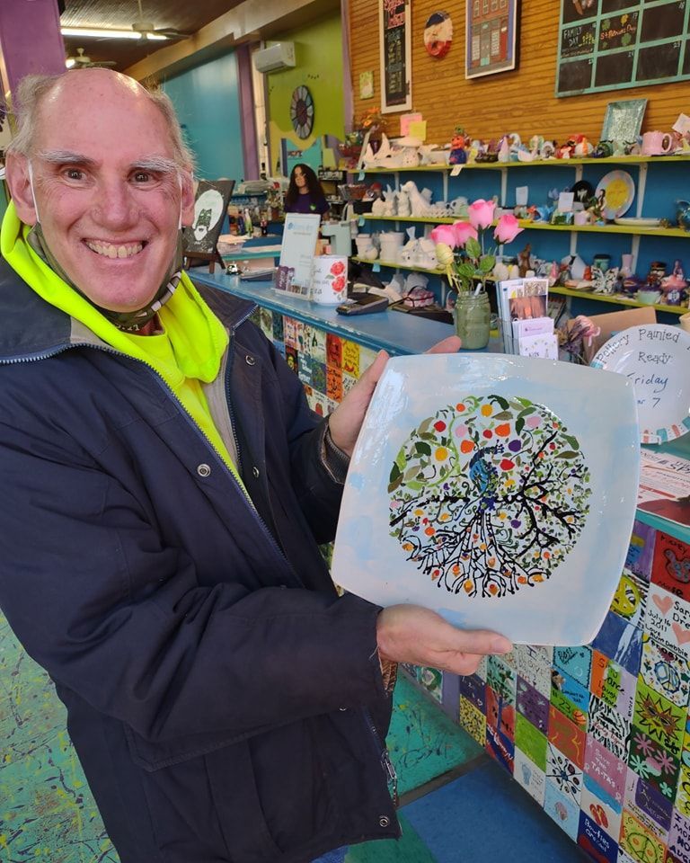 Man holding a decorated square plate with a blue bird design, smiling, inside a shop with art supplies.