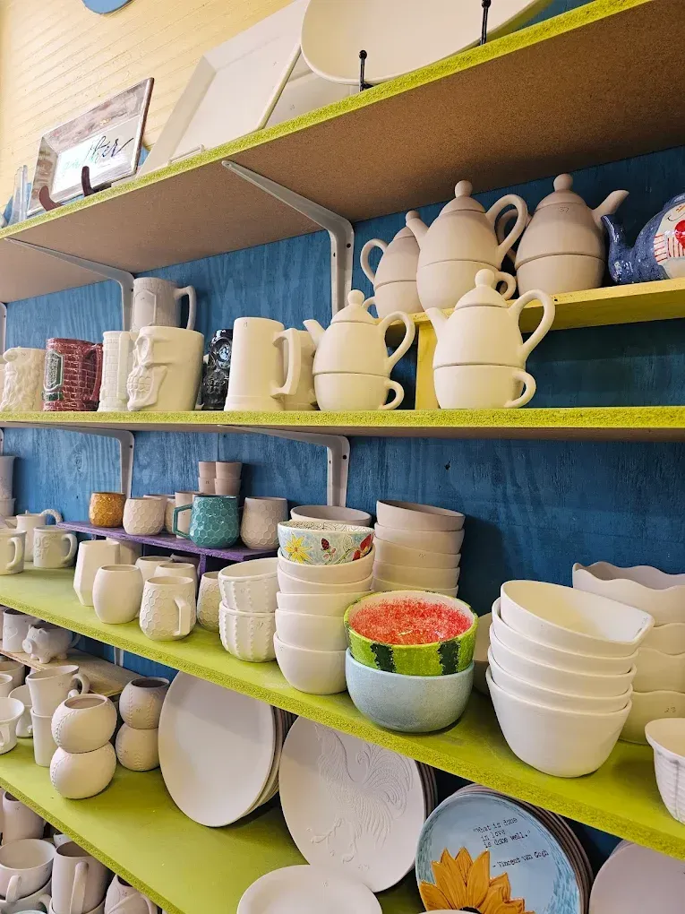 Shelves of unpainted ceramic pottery: mugs, teapots, bowls, plates, and a watermelon-decorated bowl.