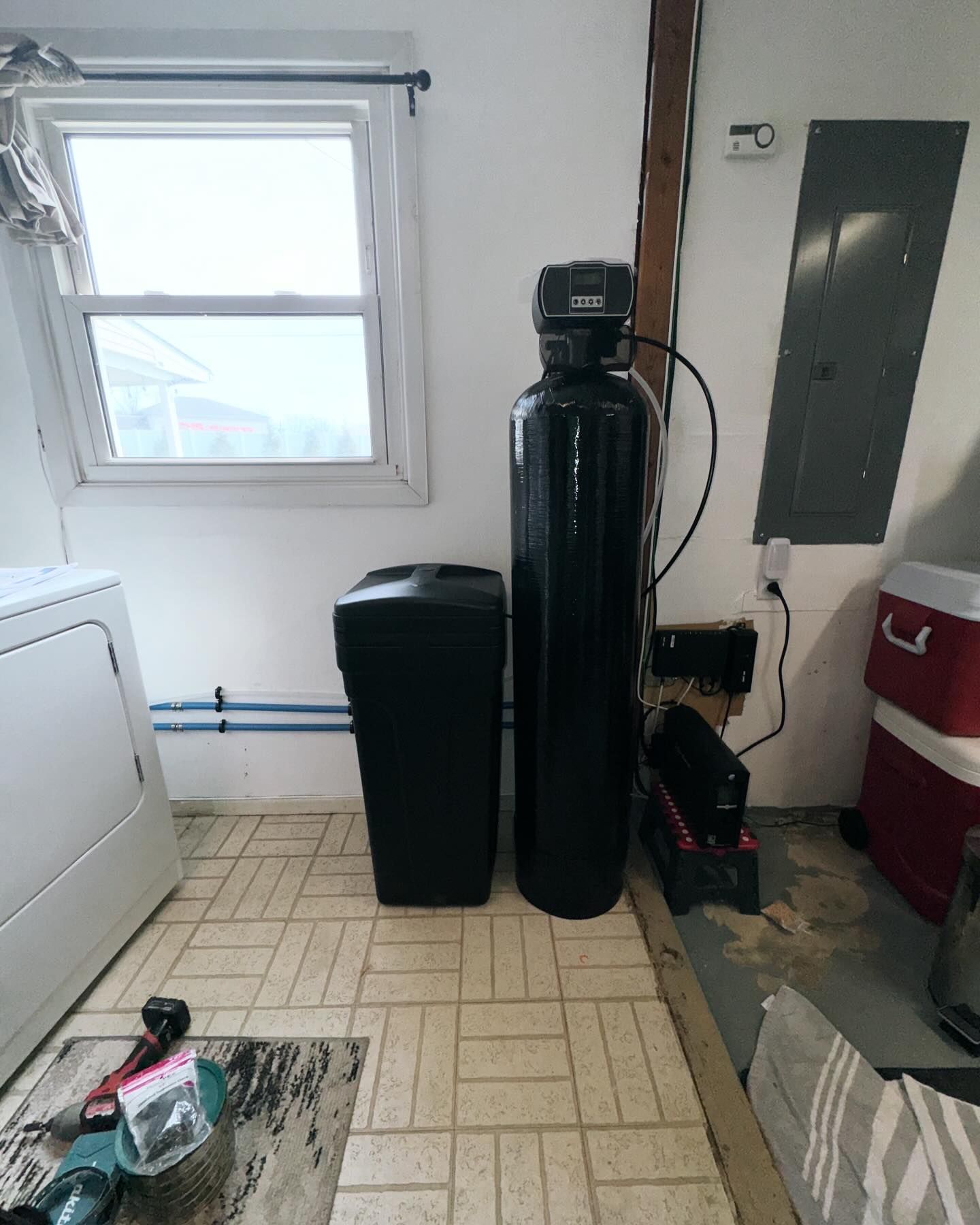 Water softener system in a laundry room, with a tall black tank, salt tank, and window.