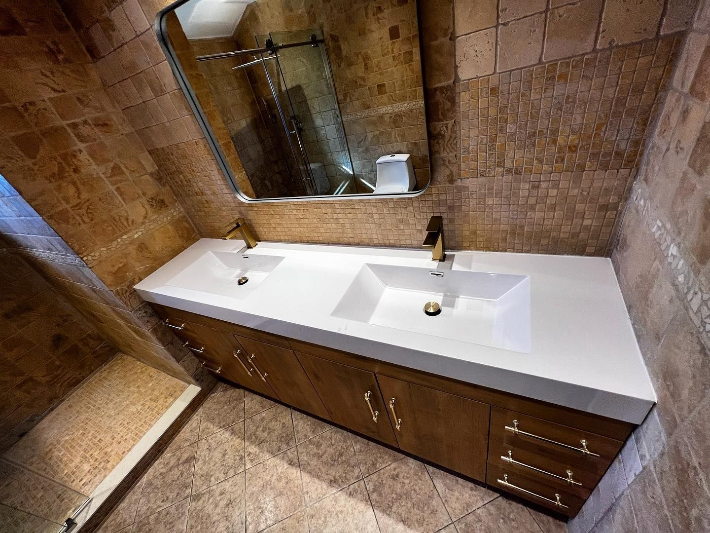 Double sink vanity with gold fixtures, white countertop, brown cabinet, and stone tile walls.