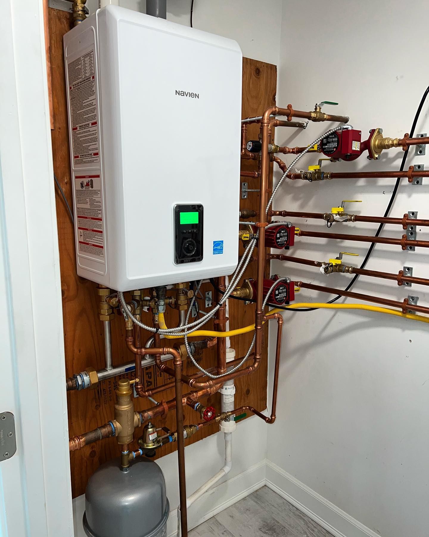 White wall-mounted water heater with copper pipes on a wooden panel. Includes expansion tank and control panel.