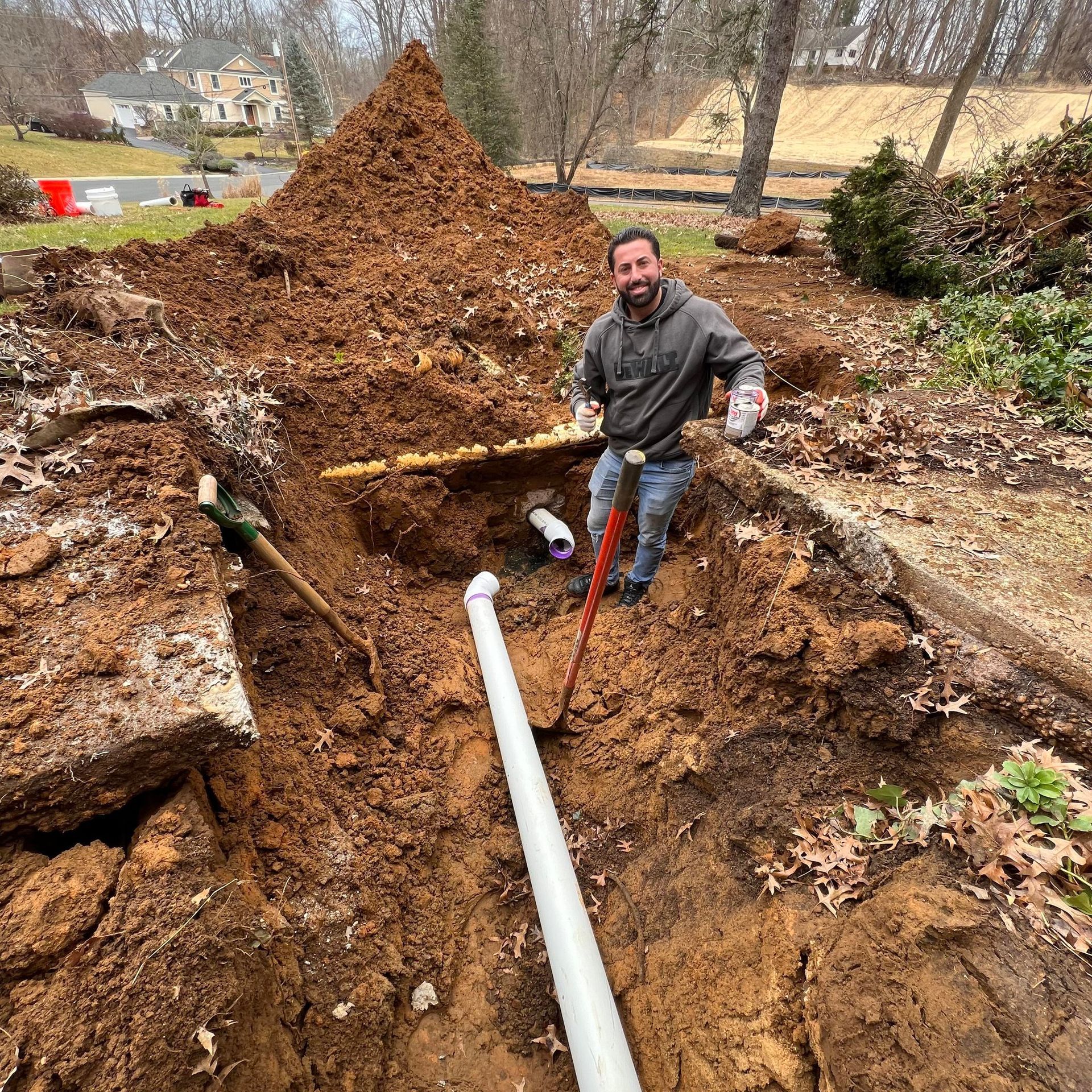 Man in a trench holding tools, smiling. Large pile of dirt in the background, plumbing visible.