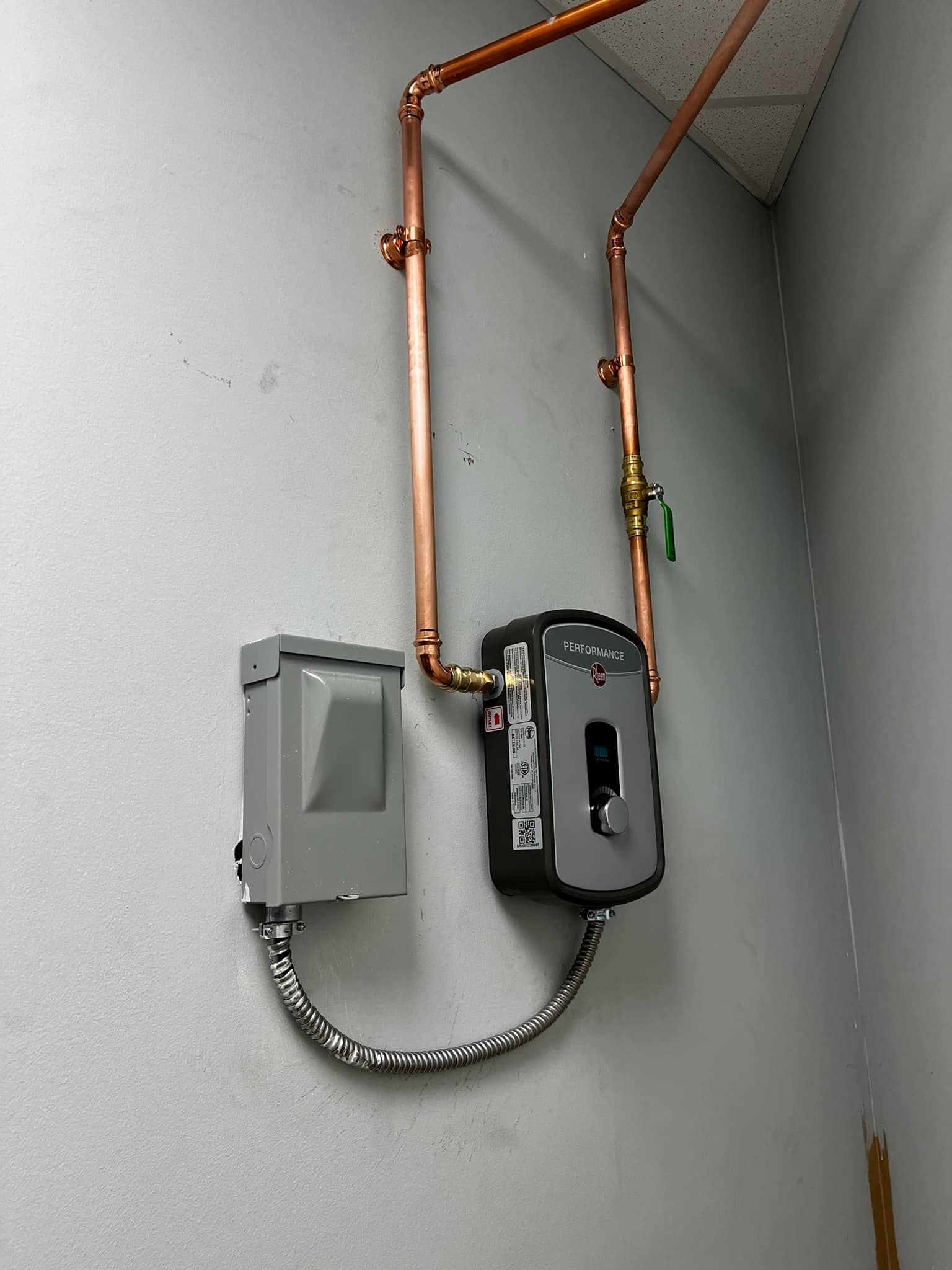 Copper pipes connect to a gray electrical box and water heater on a light gray wall.