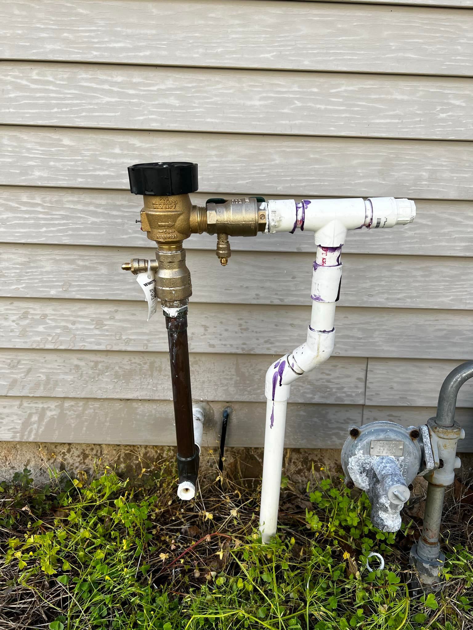 Water pipes attached to a house with a black and brass backflow preventer and white PVC pipes.