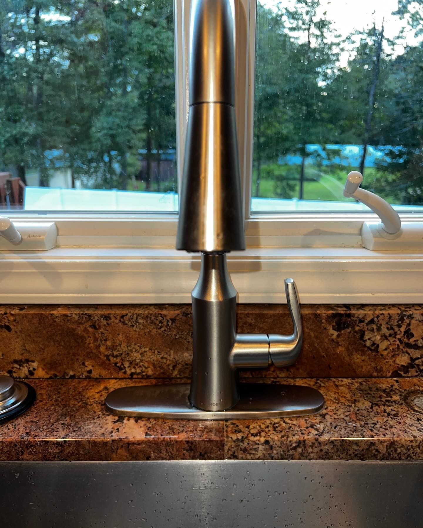 Stainless steel kitchen faucet on a speckled countertop, window in background with trees.