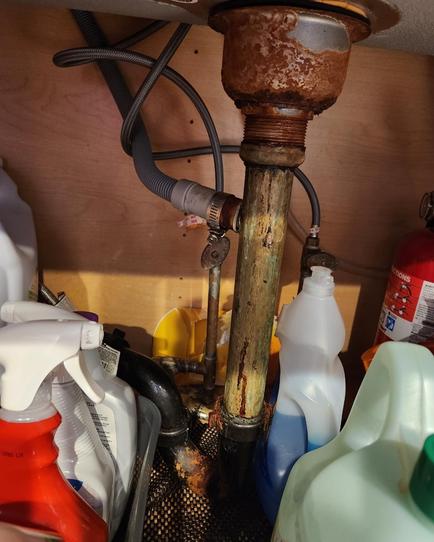 Rusty kitchen sink drain pipes under a wooden cabinet, with cleaning supplies and a fire extinguisher.