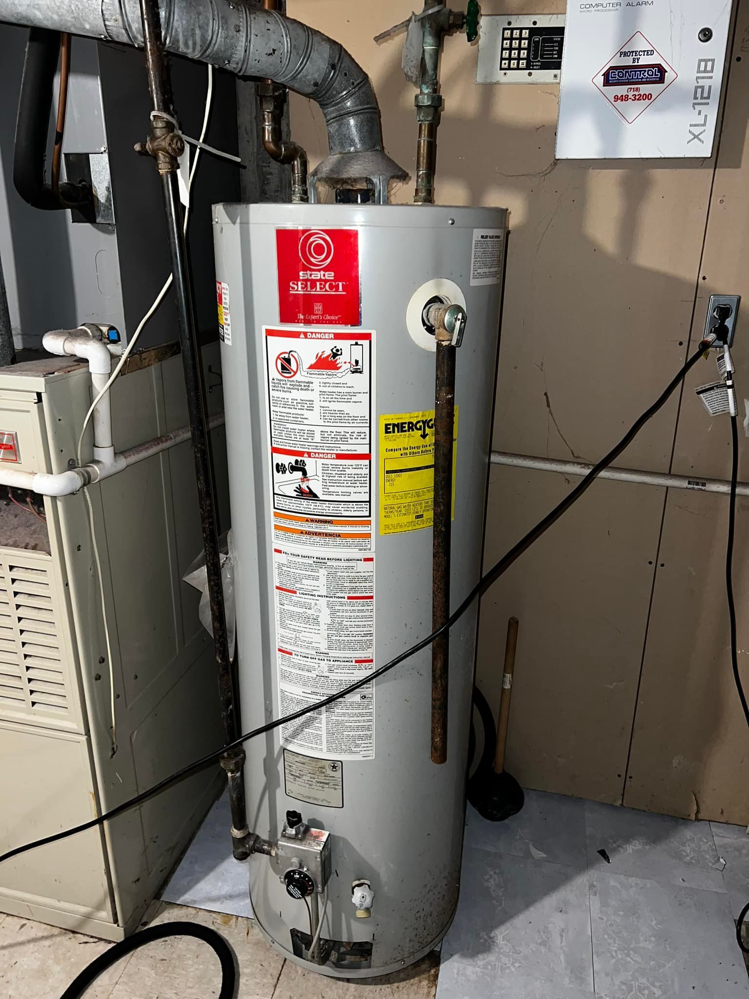 A tall, gray water heater in a basement. It has various pipes and labels attached.