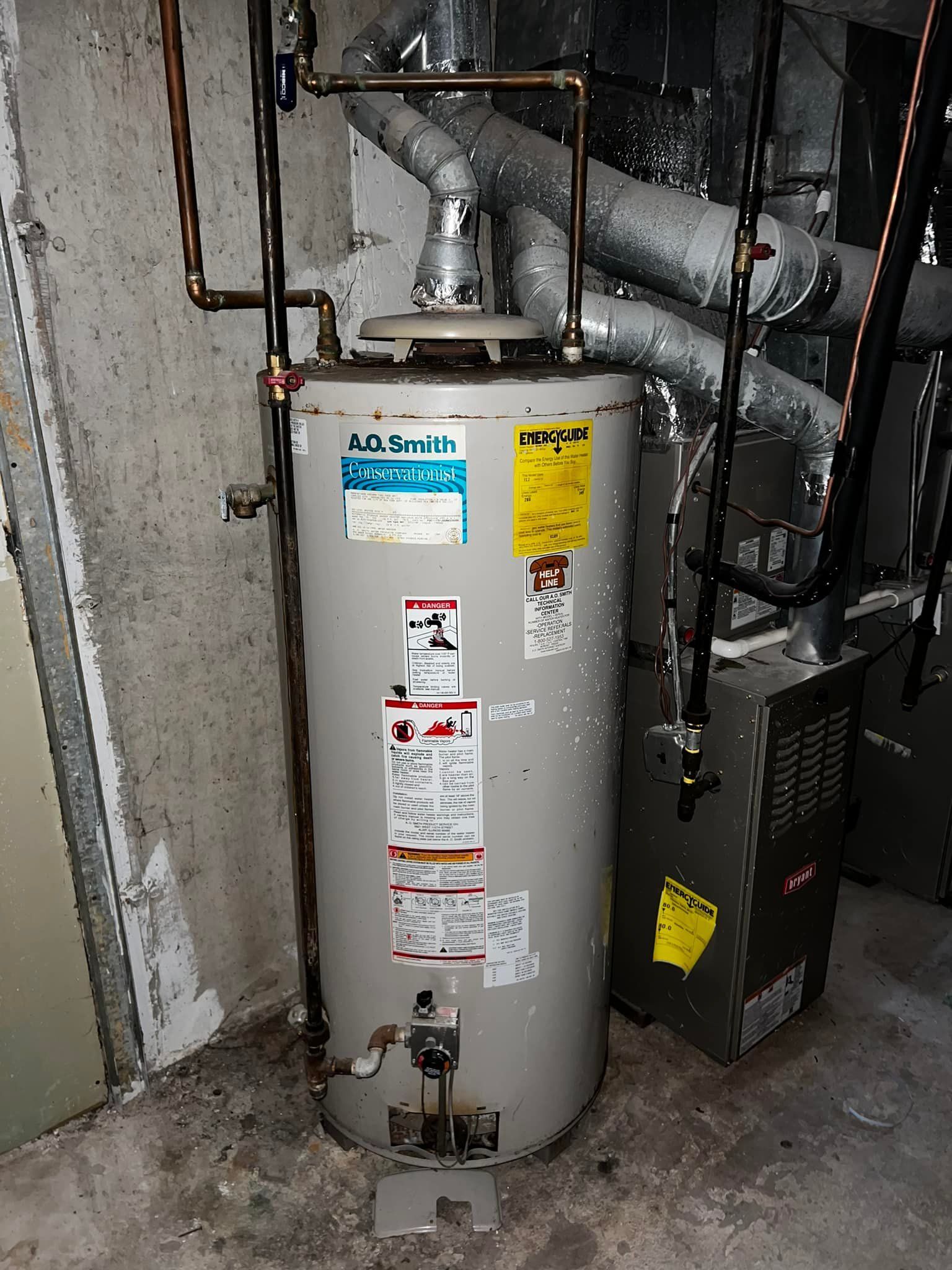 A tall, cylindrical water heater in a basement setting with various pipes and furnace nearby.