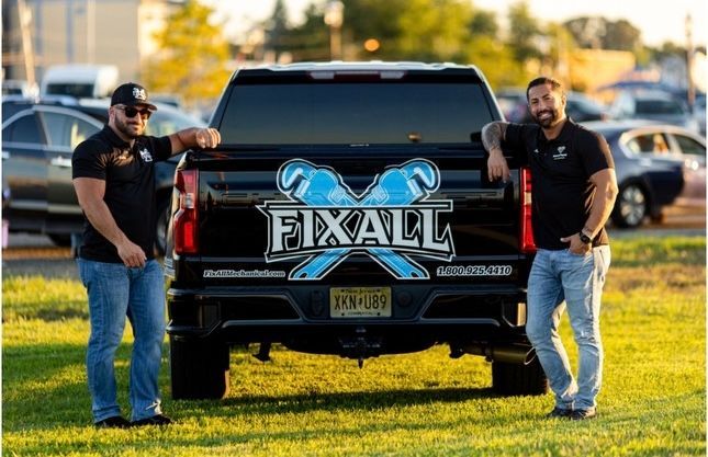 Two men lean on a black Fixall truck in a grassy area. The logo features crossed wrenches.