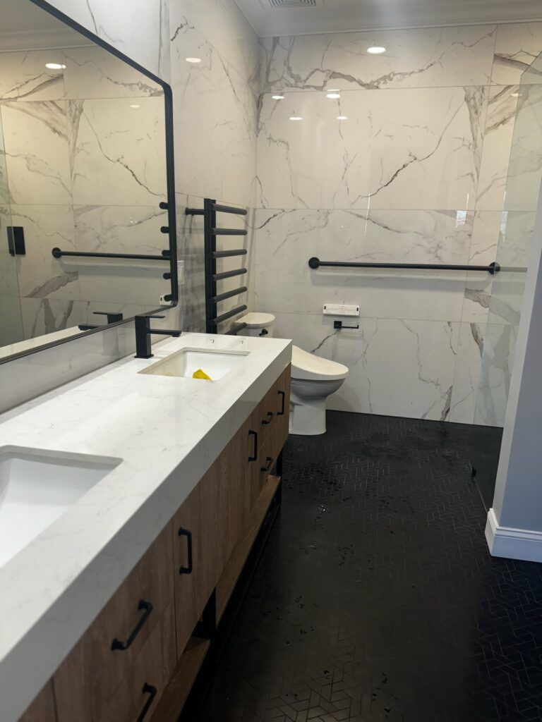 Modern bathroom with marble walls, black floor, double vanity, and a toilet with grab bars.