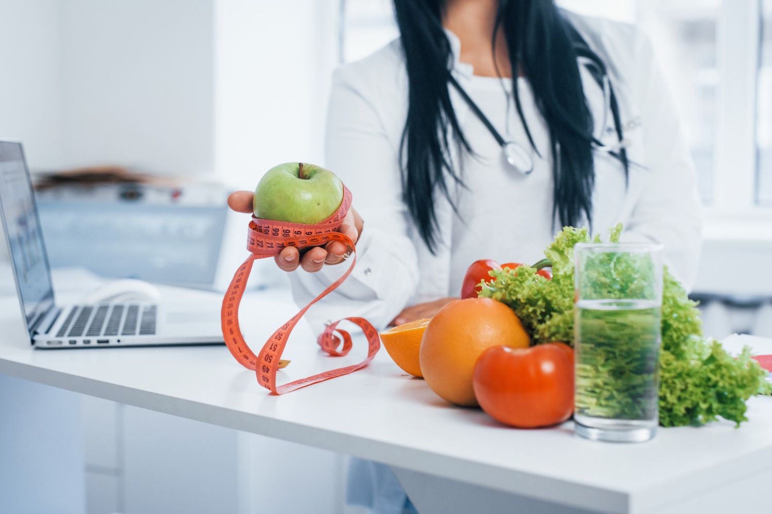 Dietitian holding apple wrapped with measuring tape, table with fruit, vegetables, and glass of water.
