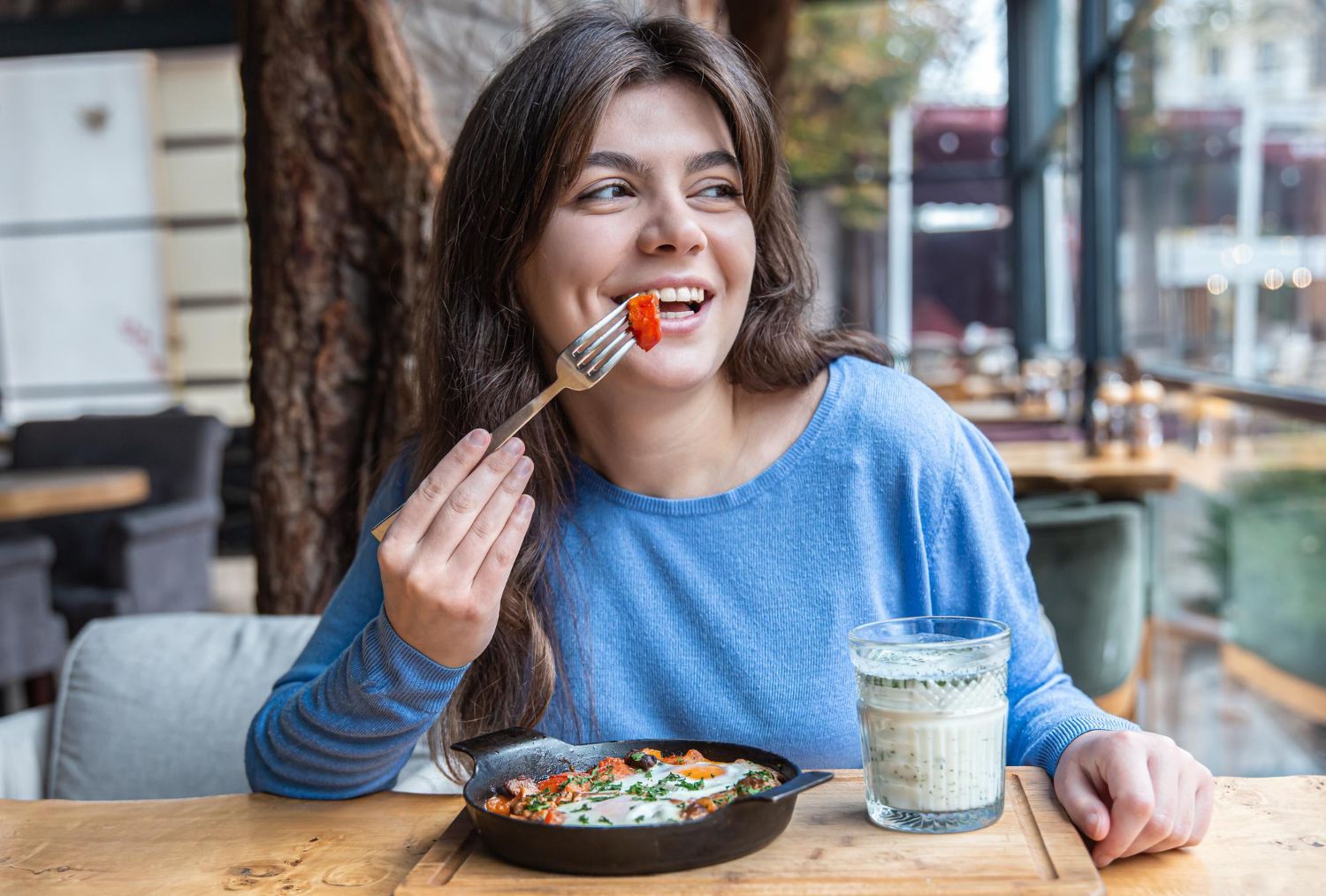 Woman eating a meal at an outdoor cafe, smiling beside a drink and bowl on a wooden table.
