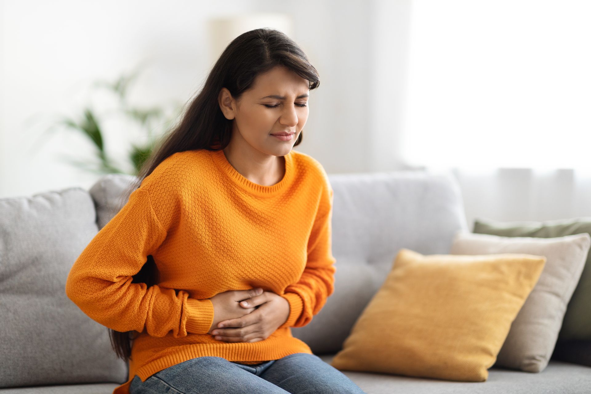 Woman with stomach pain, sitting on a sofa in a living room, holding her abdomen, wearing an orange sweater.