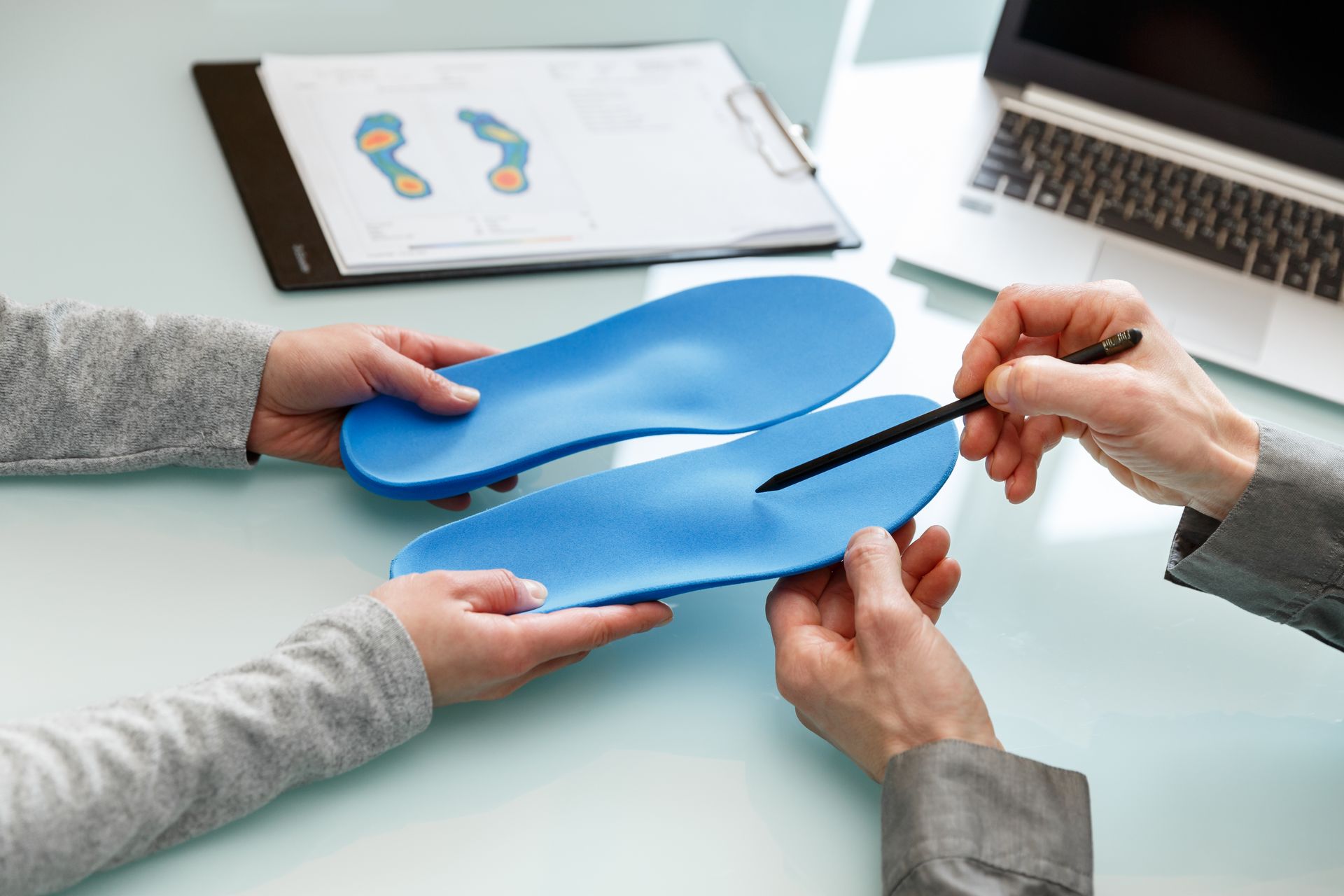 Hands examining blue orthotics, a foot scan, and a laptop on a table.