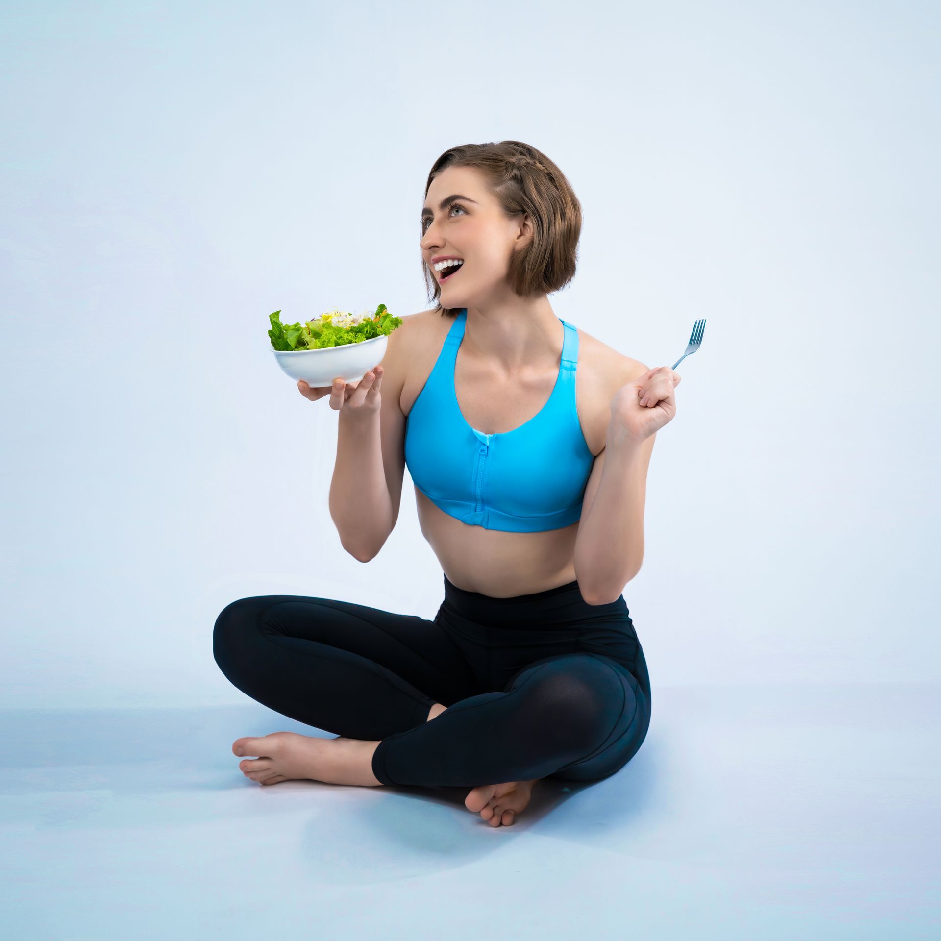 A woman is sitting on the floor eating a salad with a fork.