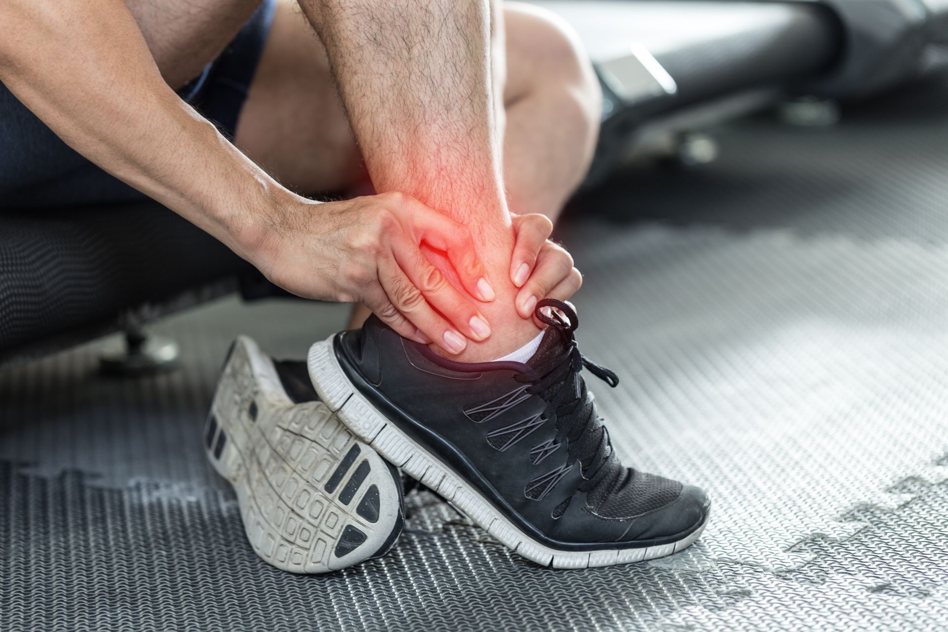 Man holding injured ankle, pain highlighted in red, wearing athletic shoes. Gym setting.