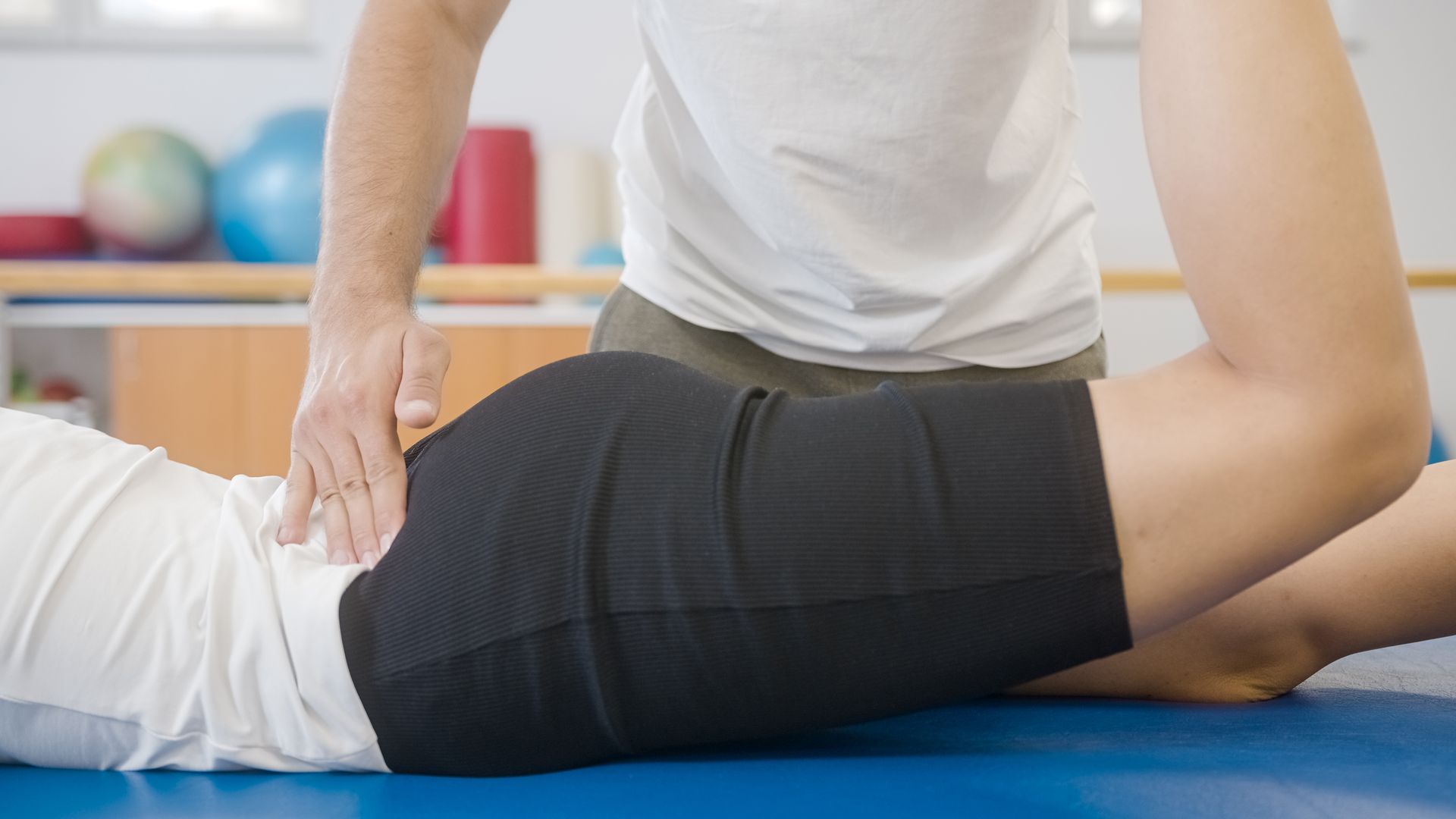 Physiotherapist assisting patient with hip exercise. Hands on lower back and leg. Blue mat, white shirt.